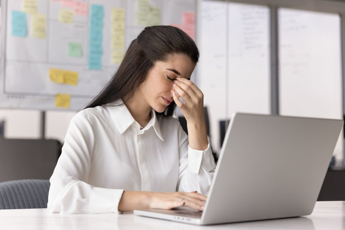 Femme au bureau se pinçant la racine du nez devant son ordinateur portable, signe de fatigue visuelle et de sécheresse oculaire